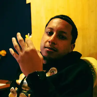 A man in a dark hoodie sitting against a yellow wall, gesturing as he talks