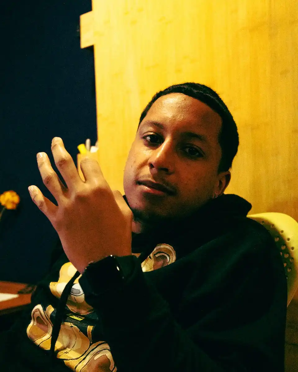 A man in a dark hoodie sitting against a yellow wall, gesturing as he talks