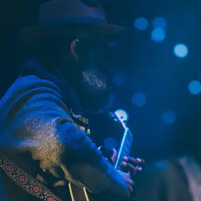 A bearded guitarist in a hat playing under blue stage lights