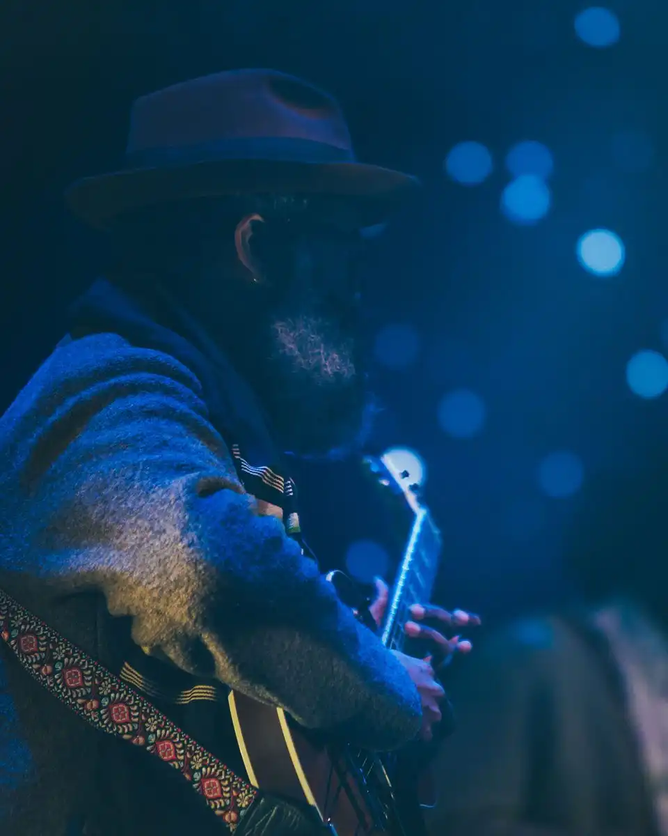 A bearded guitarist in a hat playing under blue stage lights