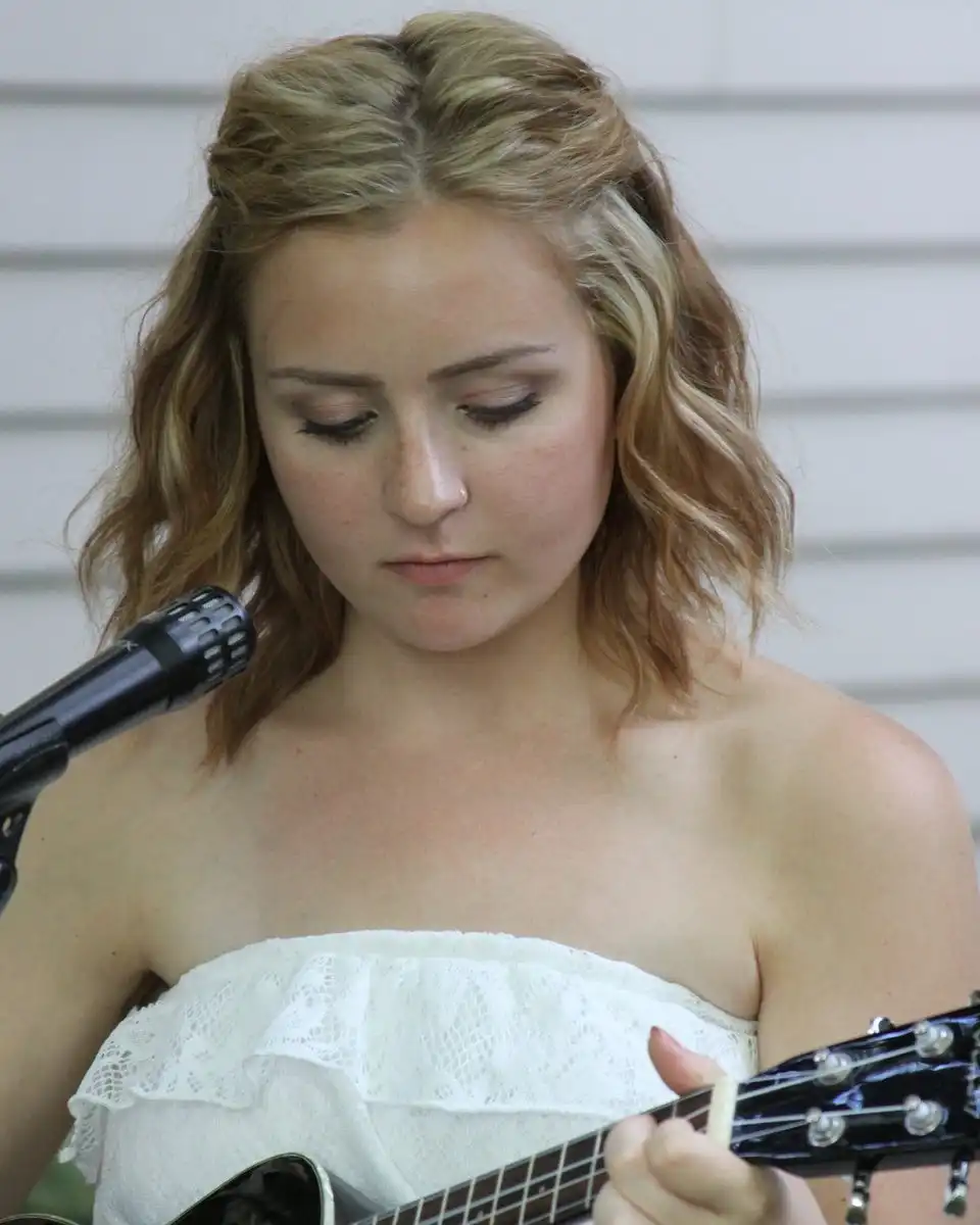 A woman with wavy hair playing a small guitar beside a microphone, looking down