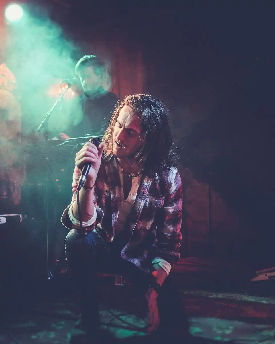 A long-haired singer in a plaid shirt crouched at a microphone in green stage haze