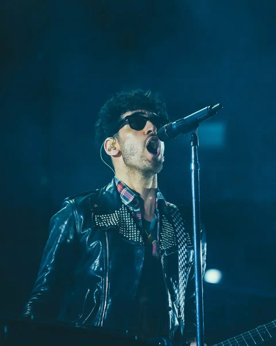 A singer in sunglasses and a studded leather jacket at a microphone under blue stage light