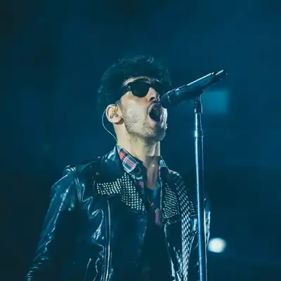 A singer in sunglasses and a studded leather jacket at a microphone under blue stage light