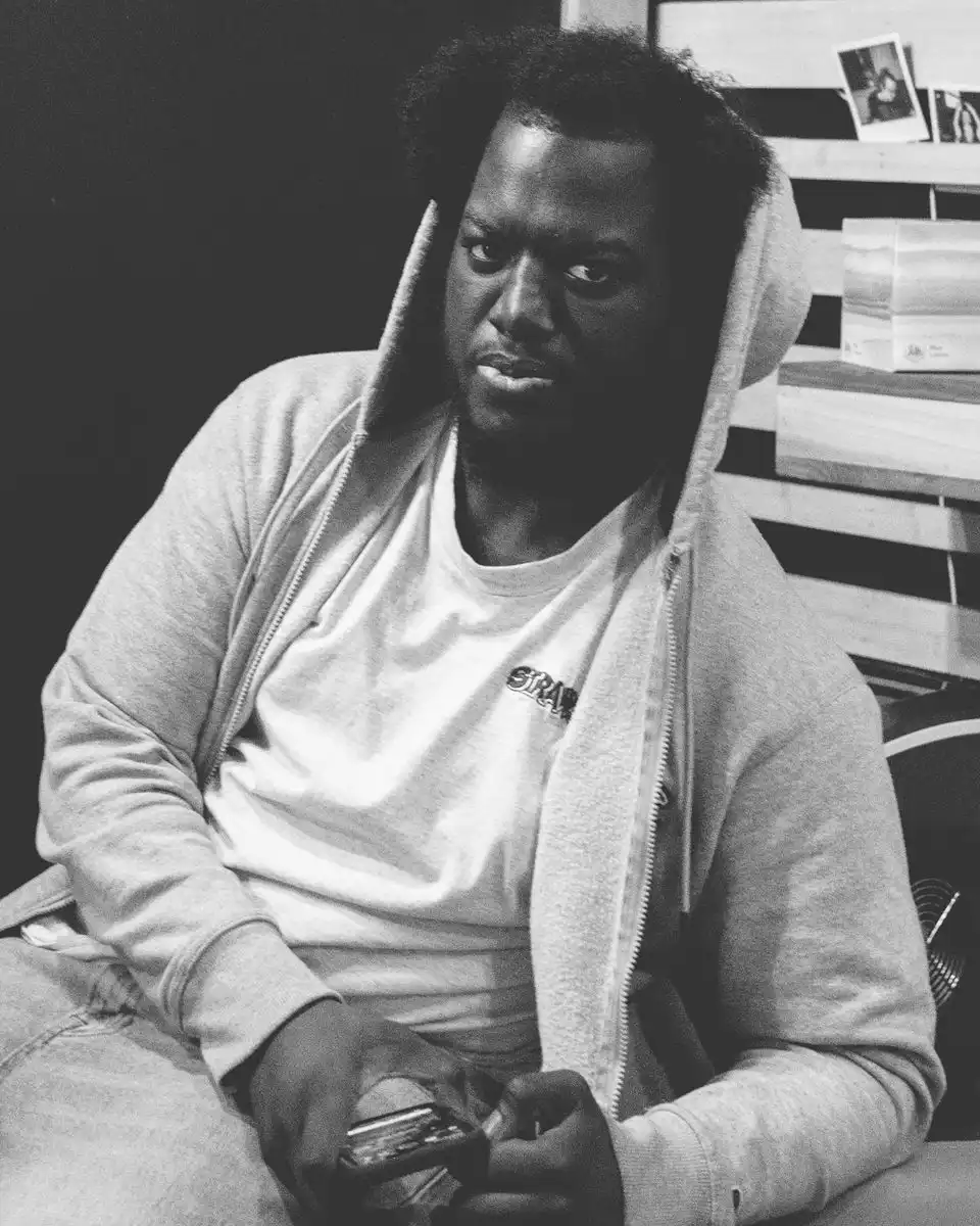 Black-and-white portrait of a man in a grey hoodie sitting on a stool in a studio